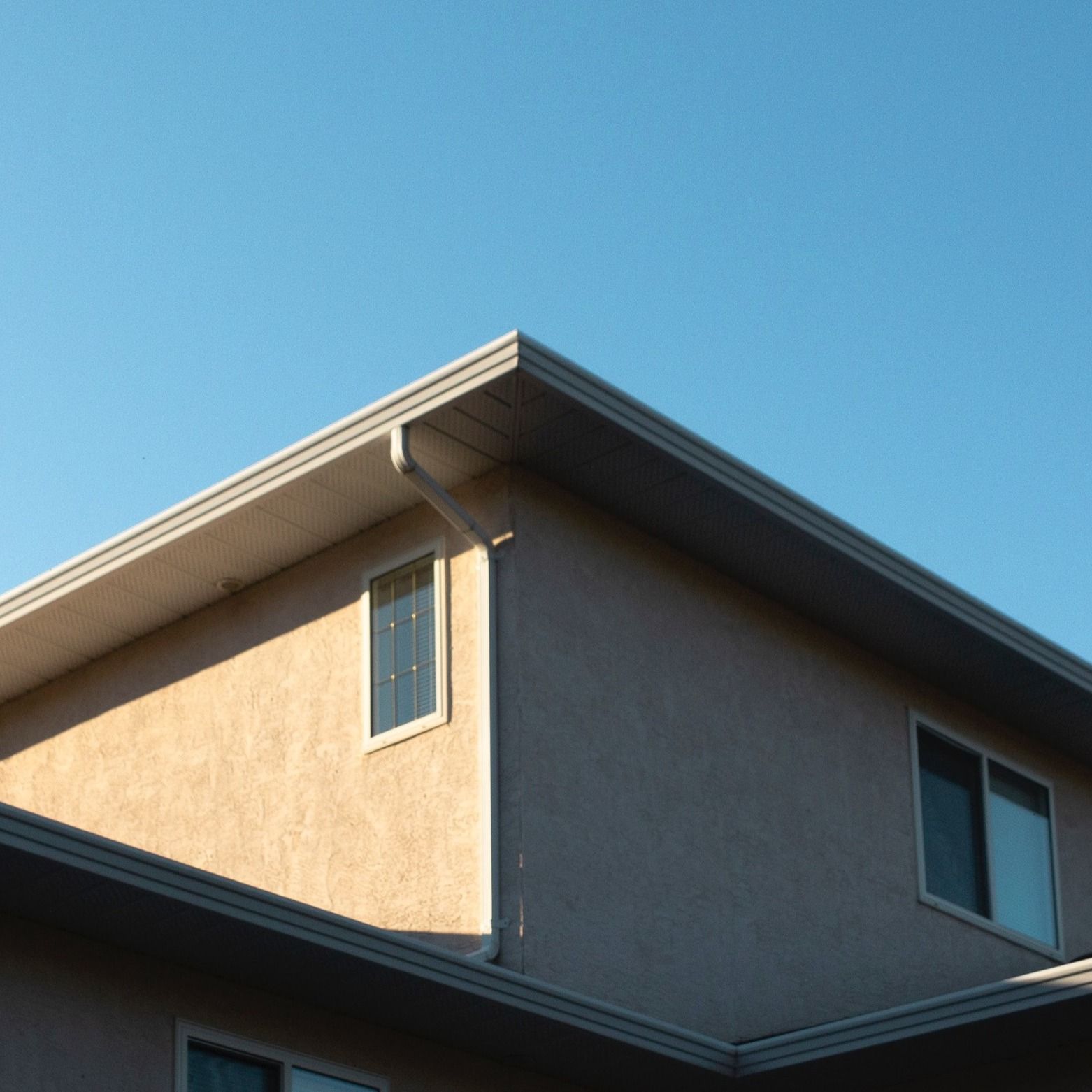 Low-angle shot of a tan-colored house with a roof overhang, gutter, and downspout against a clear blue sky.
