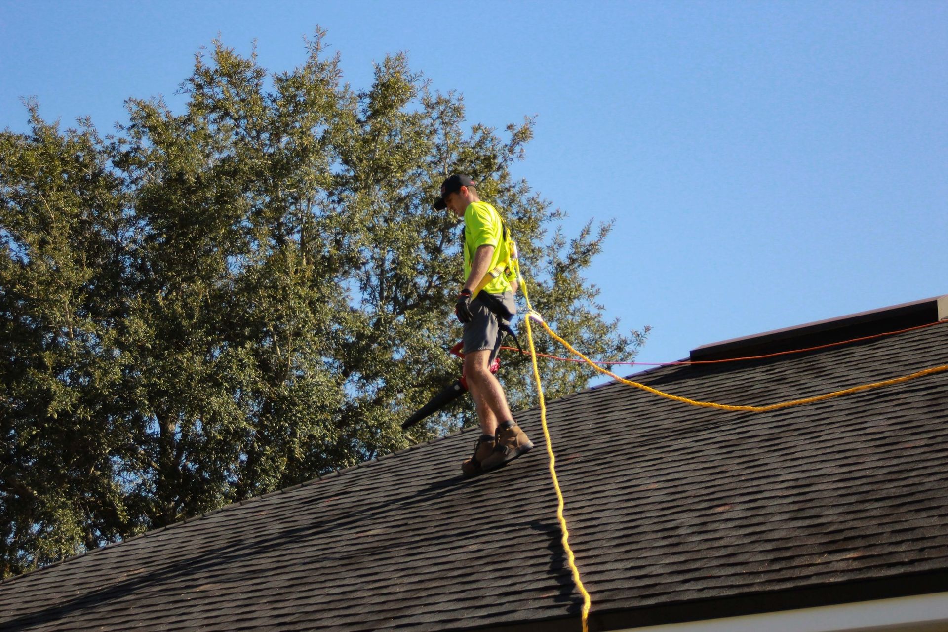 A person wearing a neon yellow safety vest and a harness is using a leaf blower while balanced on a sloped shingled roof.