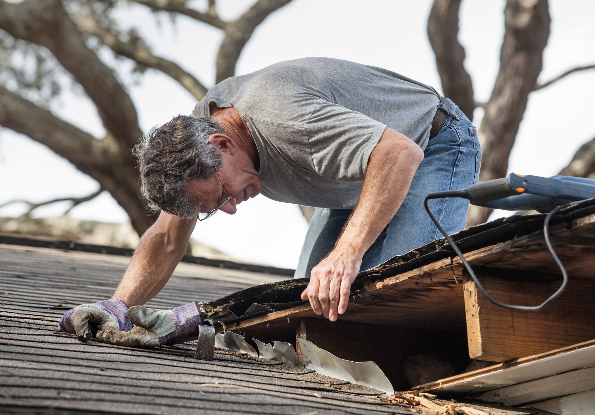 A person in a gray shirt kneels on a roof, working on removing or repairing damaged wooden roof decking.
