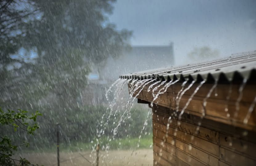 Heavy rain falls on the corrugated metal roof of a wooden building, with water streaming off the edge.