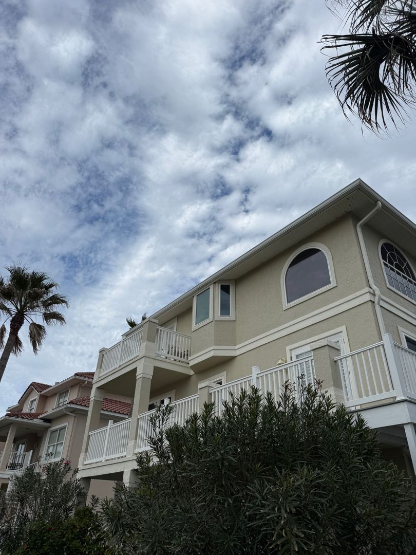 A tan, multi-story beach house with balconies and arched windows under a cloudy blue sky, framed by palm trees.