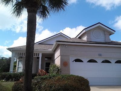 A light-colored, single-story suburban house with a two-car garage and a palm tree in the foreground under a blue sky.