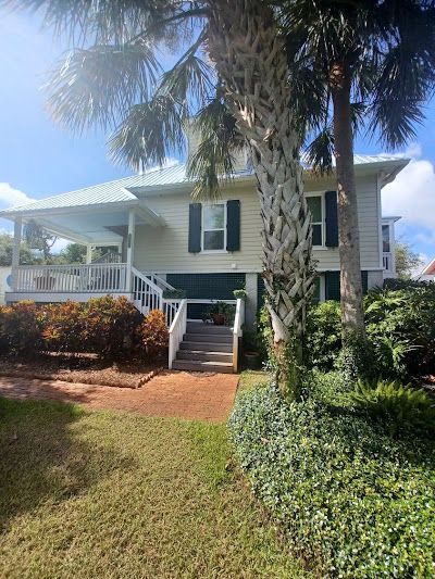 A white house with a covered porch and wooden steps, surrounded by green landscaping and a large palm tree under a blue sky.