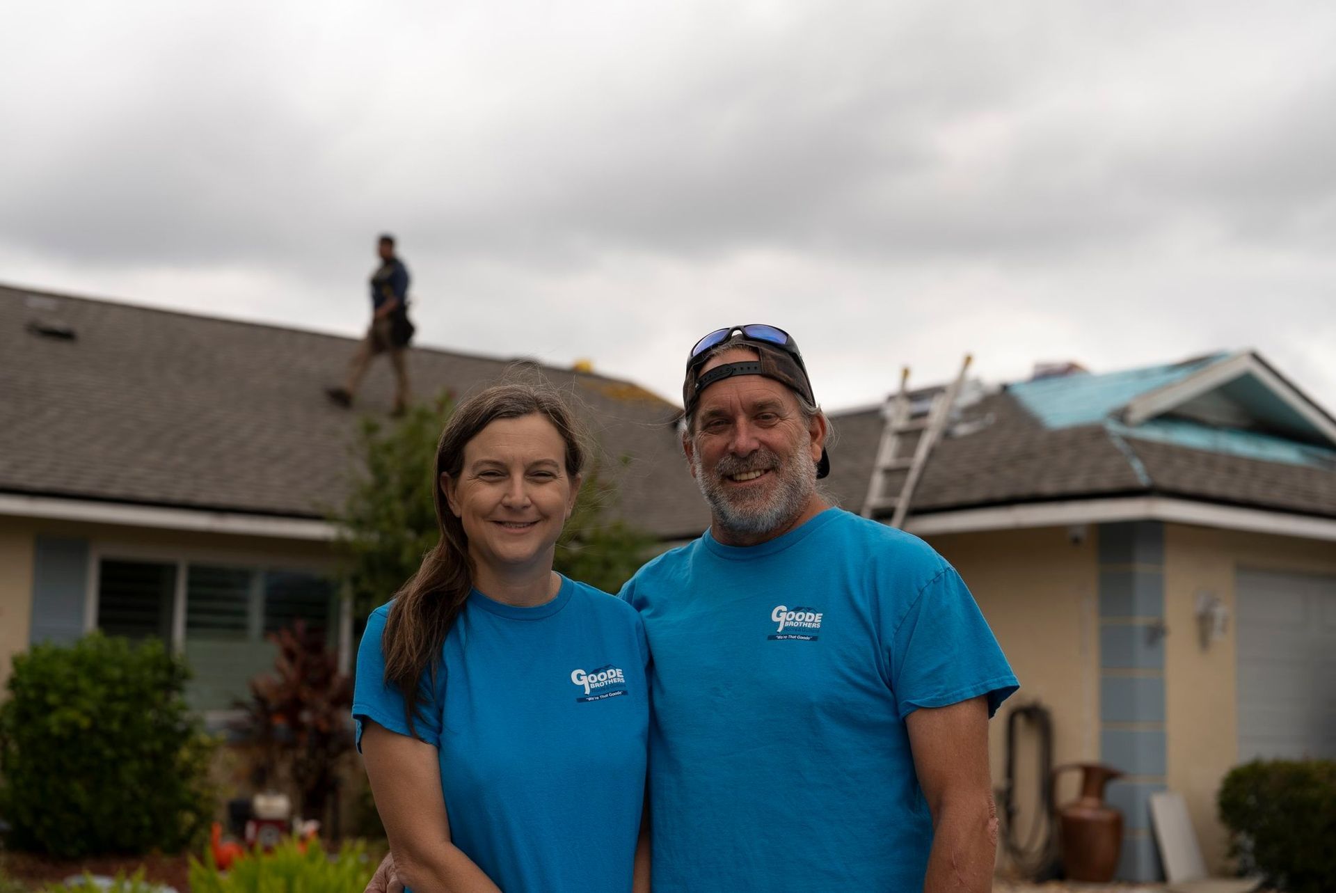 A smiling couple in matching blue shirts stands in front of a house with a person working on the roof in the background.