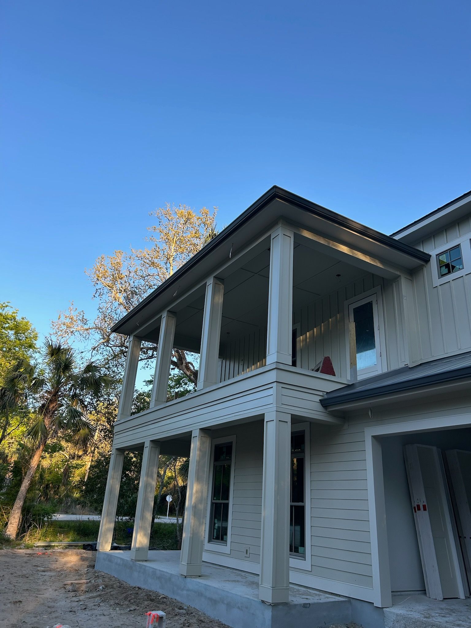 A two-story house under construction with a wraparound porch, white siding, and a clear blue sky in the background.