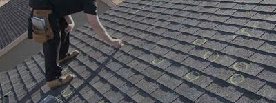 A worker wearing a tool belt inspects a residential roof marked with circles, likely indicating hail or storm damage.