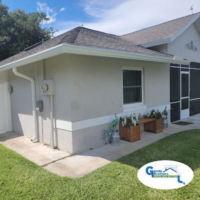 A light-colored house corner with a white gutter, a small window, two wooden planters, and a branding logo in the corner.