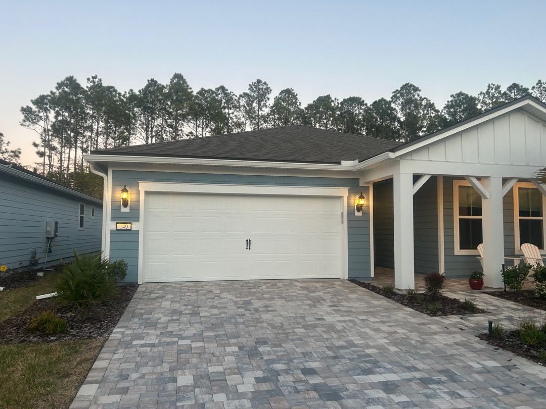 Blue single-story house with a white garage door and covered porch, featuring a stone-paver driveway at dusk.