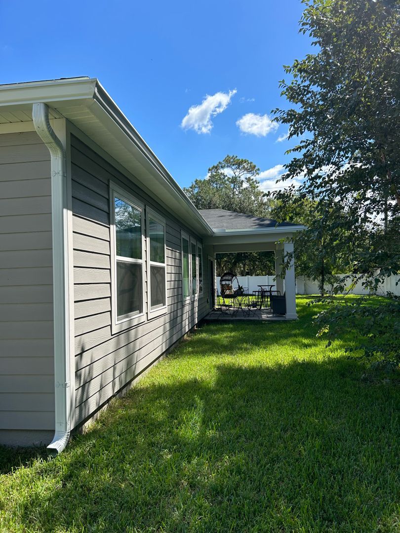 Side view of a gray-sided house with white trim, a covered patio, and a bright green lawn under a sunny blue sky.