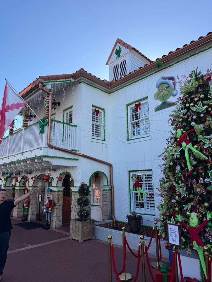 A two-story white building decorated for Christmas with a large Grinch tree, garlands, and a red and white flag.