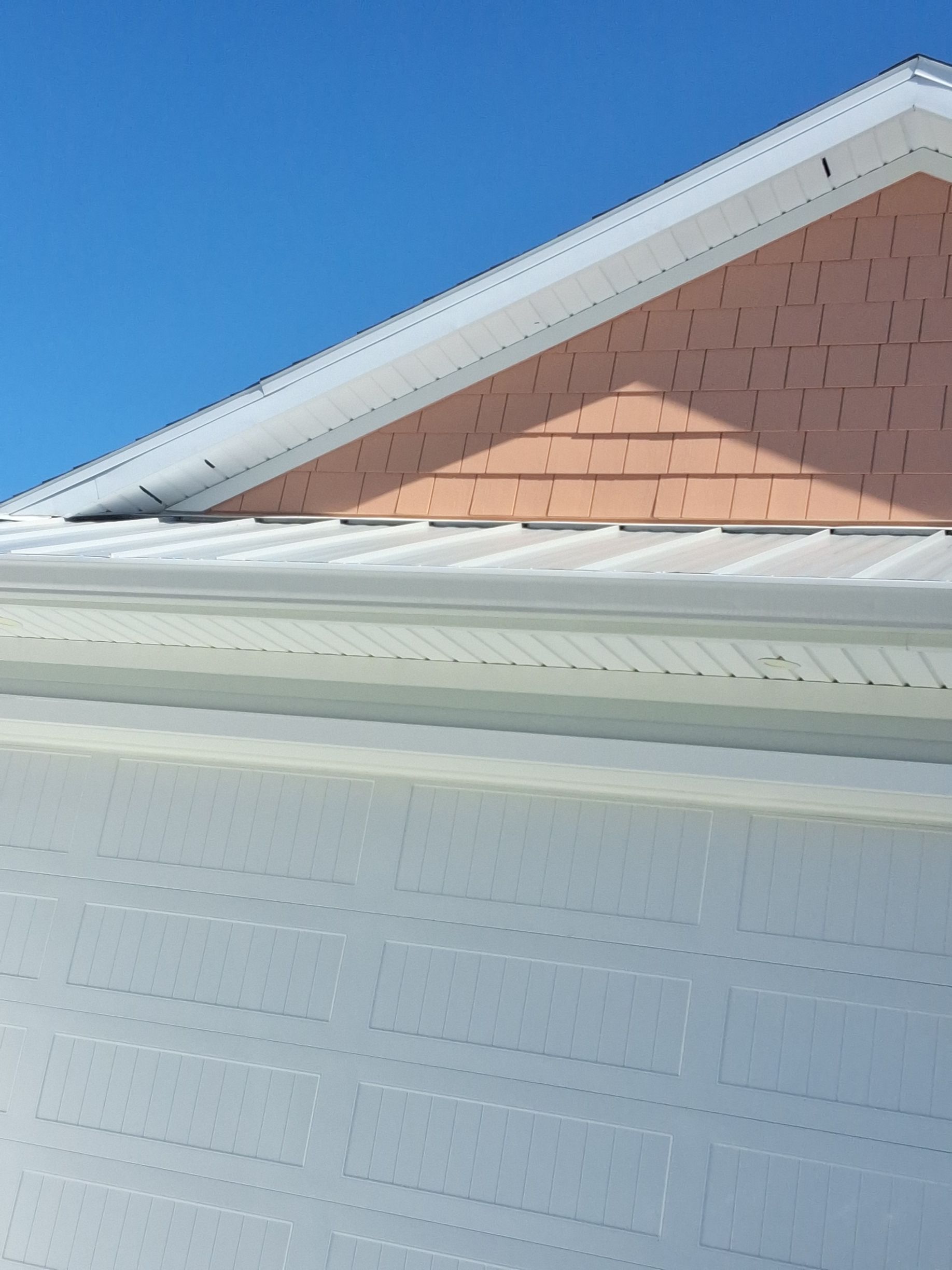 A view of a house gable with peach-colored shingles above a white metal roof and light-colored siding against a blue sky.