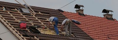 Two people work on a roof, installing brown tiles on a wooden frame next to an existing section of red tiles.