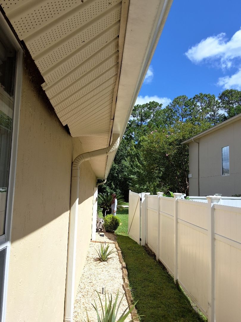 Side view of a beige house exterior with a white soffit, gutter downspout, stone landscaping, and a white privacy fence.