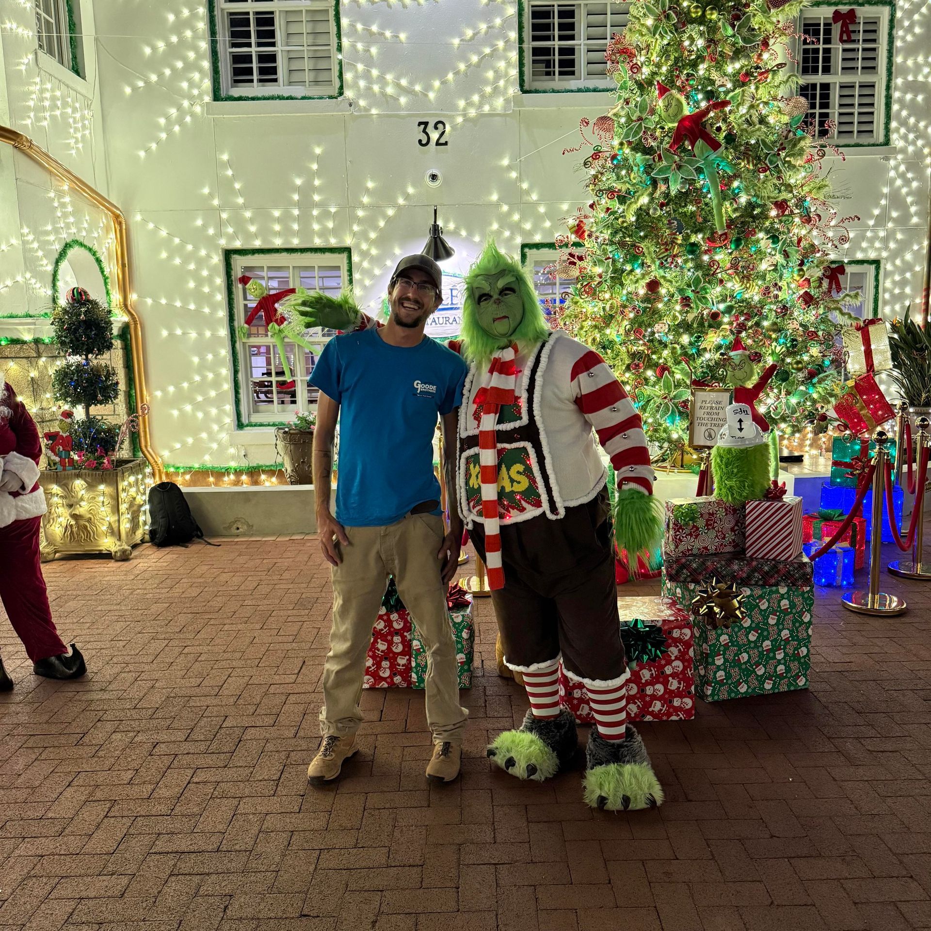 A person in a blue shirt stands next to a costumed Grinch character in front of a Christmas tree and festive lights.