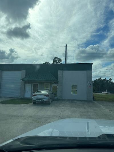 A silver sedan parked in front of a gray, one-story industrial building under a cloudy sky.