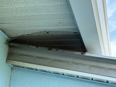 A dark, mesh-covered vent gap located under the white soffit and eaves of a light blue building exterior.