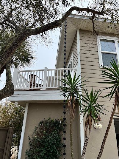 A black rain chain hangs from the roof edge of a tan two-story house with a white balcony and a palm tree nearby.