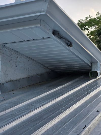 A low-angle view of a white soffit and gutter system above a sloped metal roof, showing a disconnected downspout elbow.