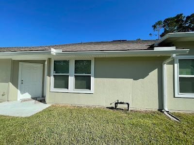 A light green house exterior with a white door, two windows, a gray shingled roof, and a green lawn under a blue sky.