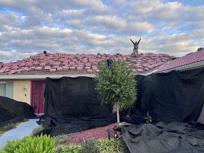 A person stands on a roof covered in scattered red tiles, surrounded by black tarps protecting the home below.