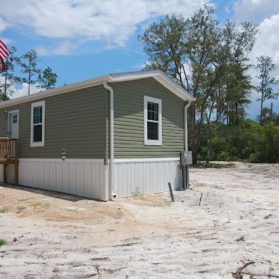 A sage green manufactured home with white skirting and a small wooden porch sits on a sandy lot under a cloudy blue sky.