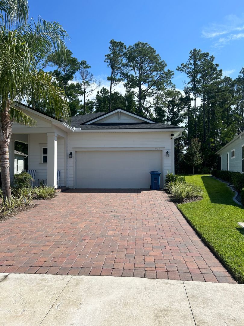 A house exterior featuring a brick paver driveway leading to a large white garage door, set under a clear blue sky.
