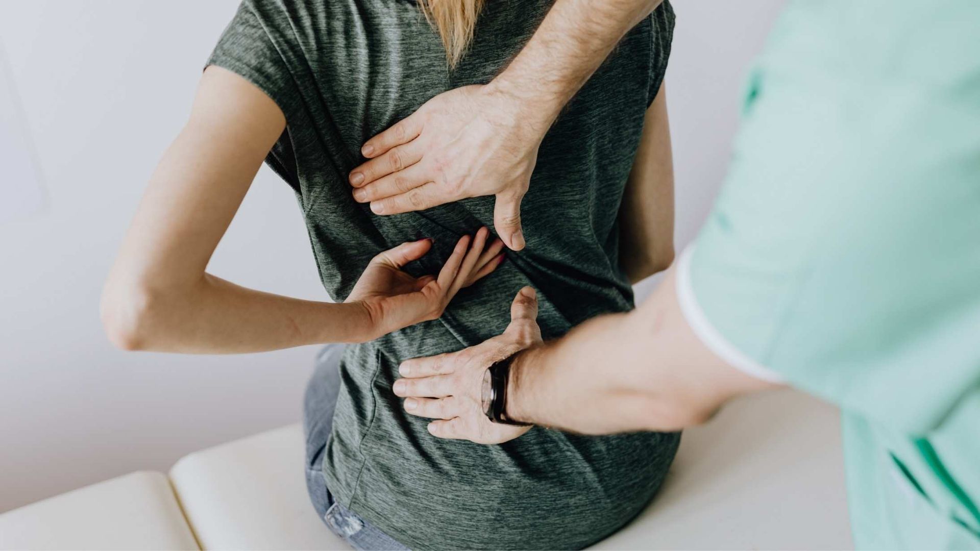 Person seated, receiving a back examination from a medical professional. Hands on lower back.