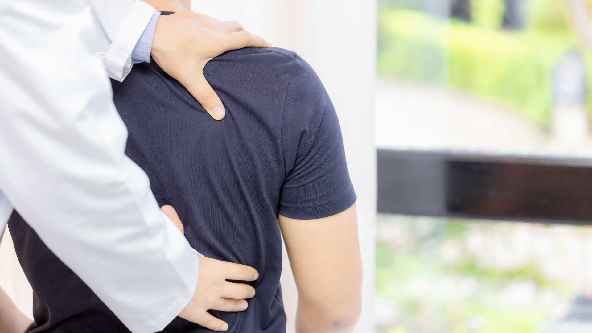 Doctor examining patient's back, hand on shoulder and lower back, inside near a window.