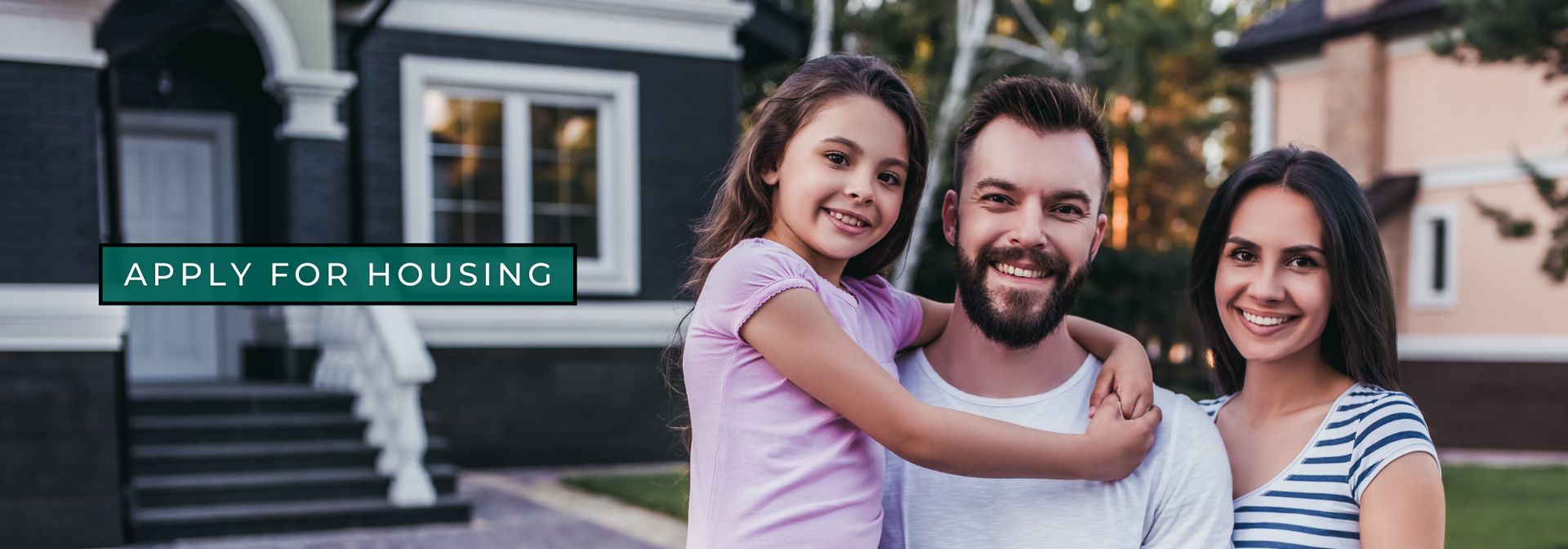 Smiling family outside a house with a green “APPLY FOR HOUSING” sign.