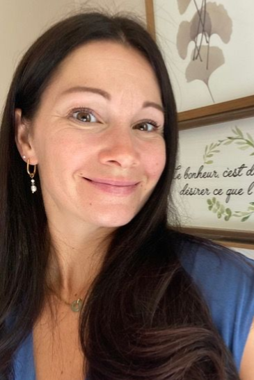 Femme souriante, longs cheveux noirs, portant une chemise bleue, tableau encadré en arrière-plan.