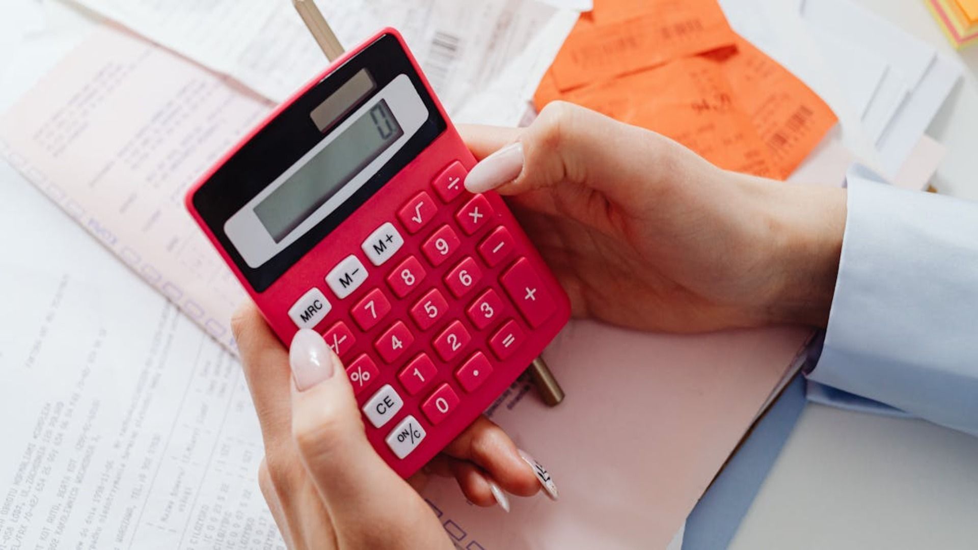 Manos usando una calculadora roja. Recibos y papeles al fondo.