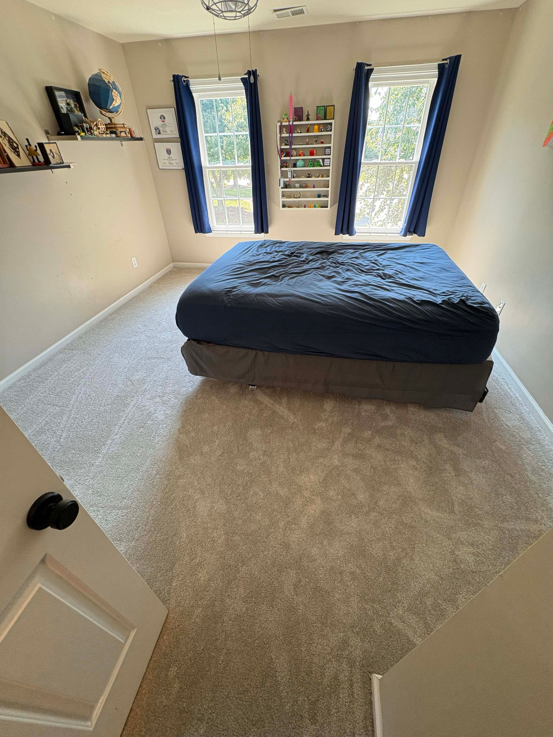 Small bedroom with bed, two windows with blue curtains, and light gray carpet.