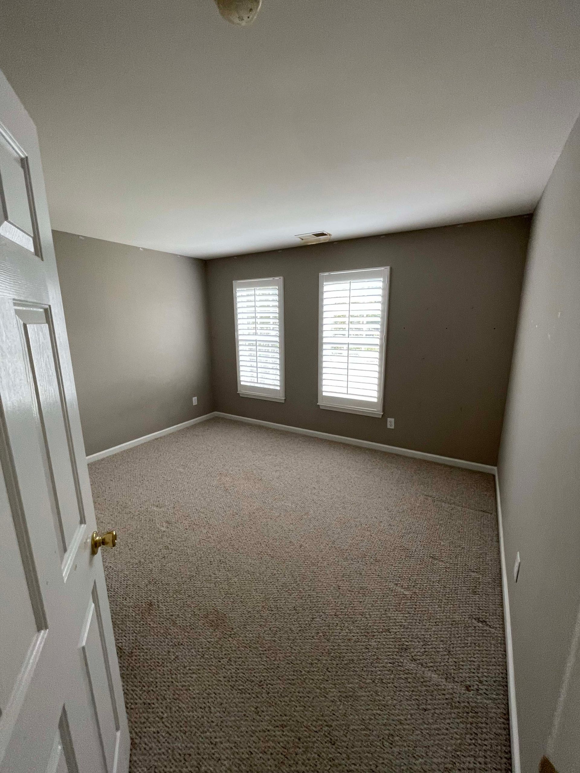 Empty bedroom with beige carpet, two windows, and neutral walls. Door partially visible.