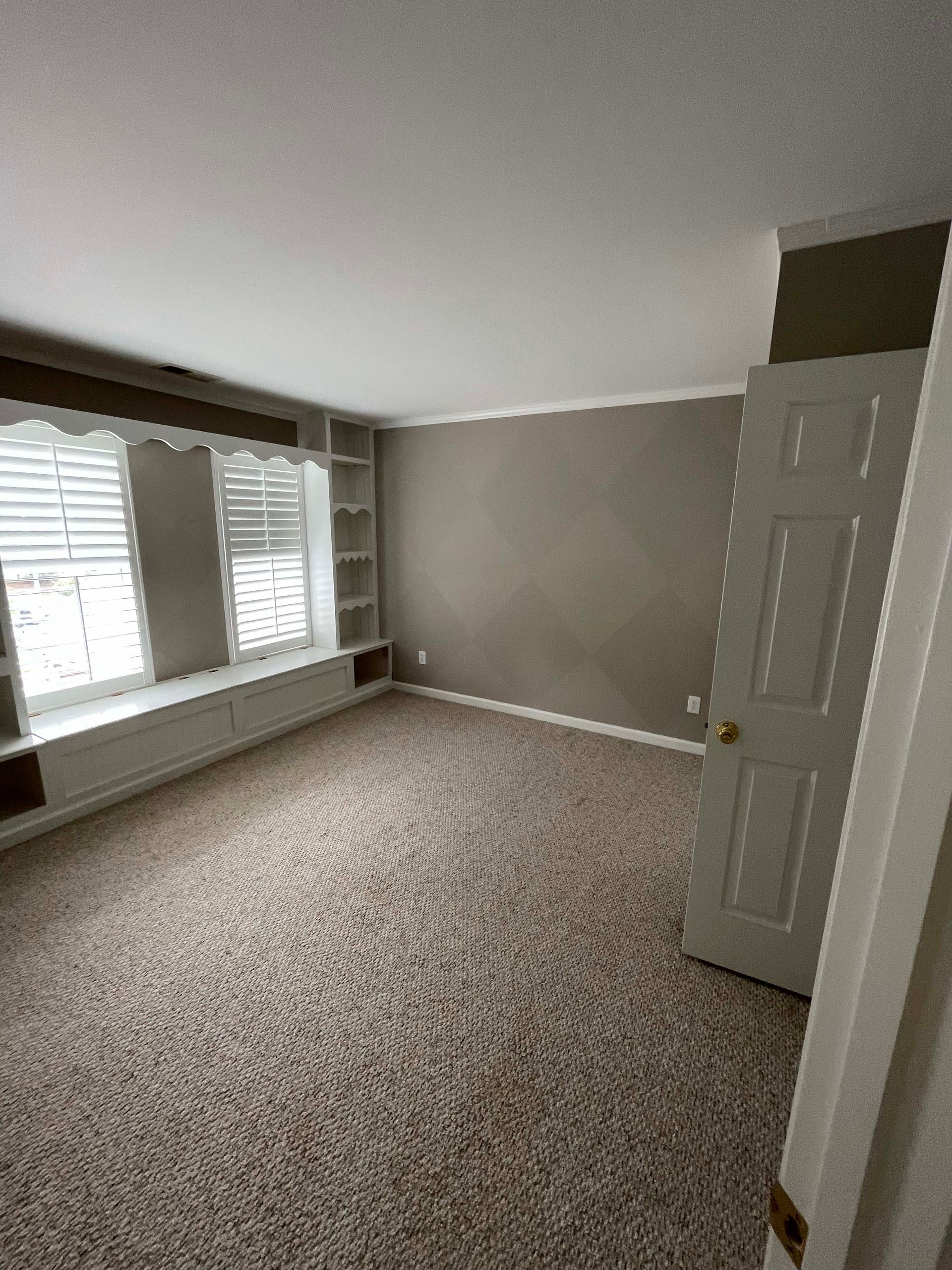Empty room with beige carpet, window seat, built-in shelving, and a closed white door.