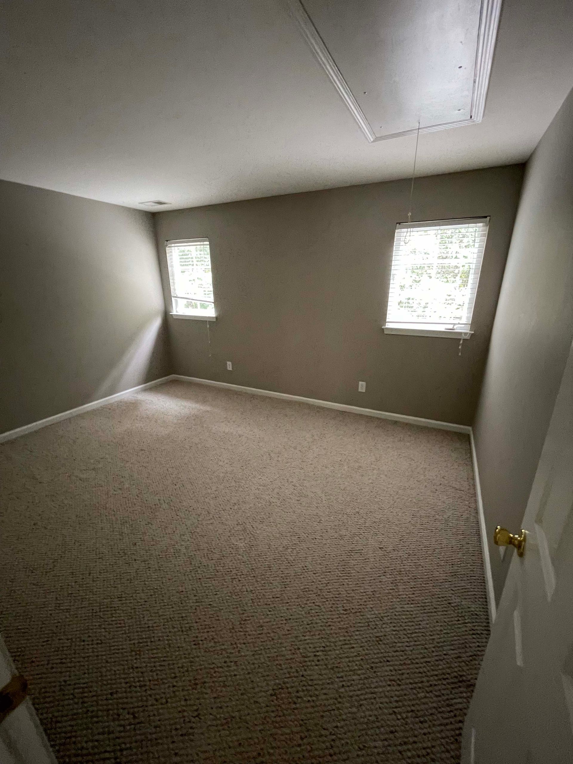 Empty room with beige walls, patterned carpet, and two windows.