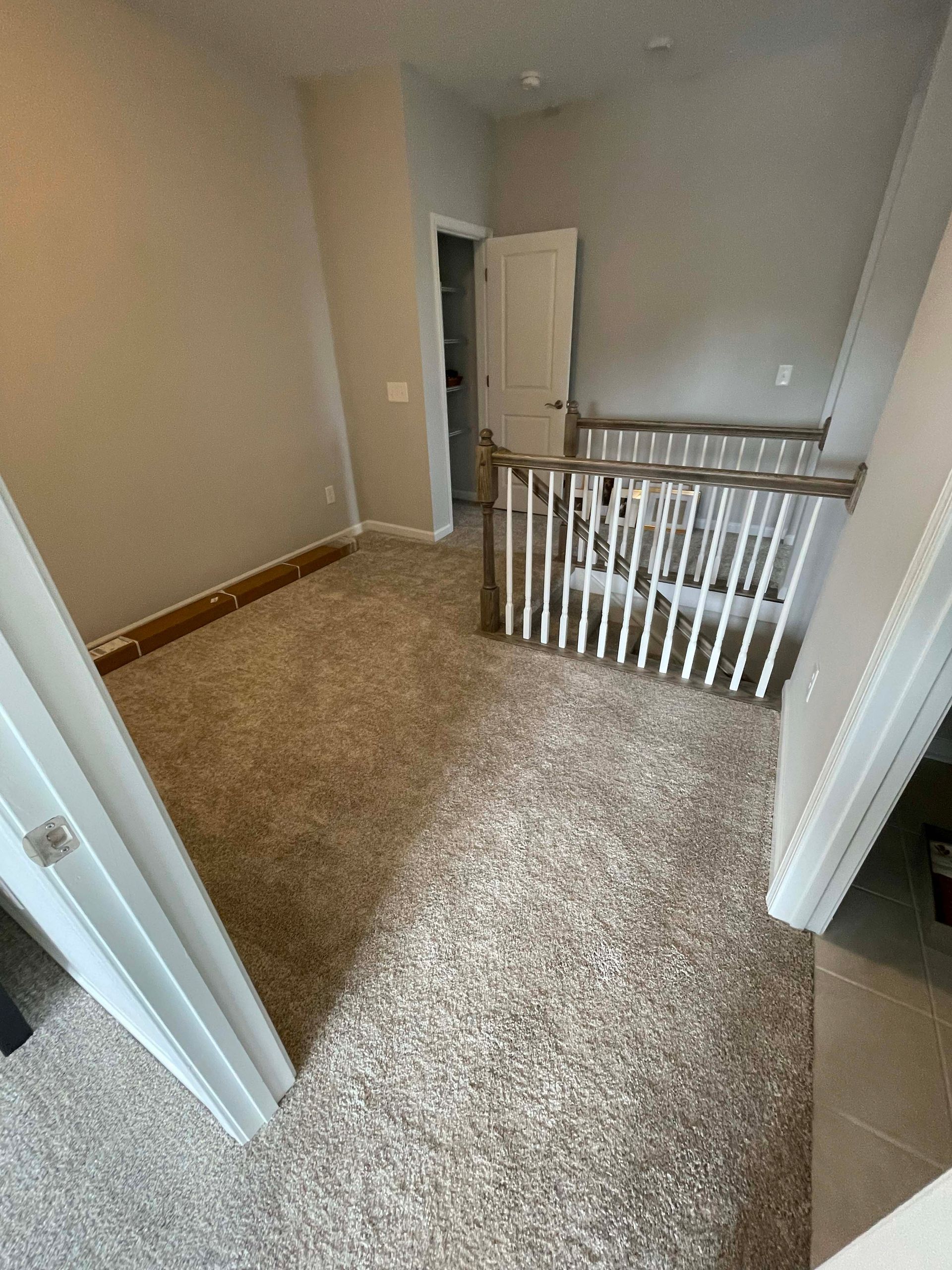 A room with beige carpet and light gray walls. A doorway and stair railing are visible.