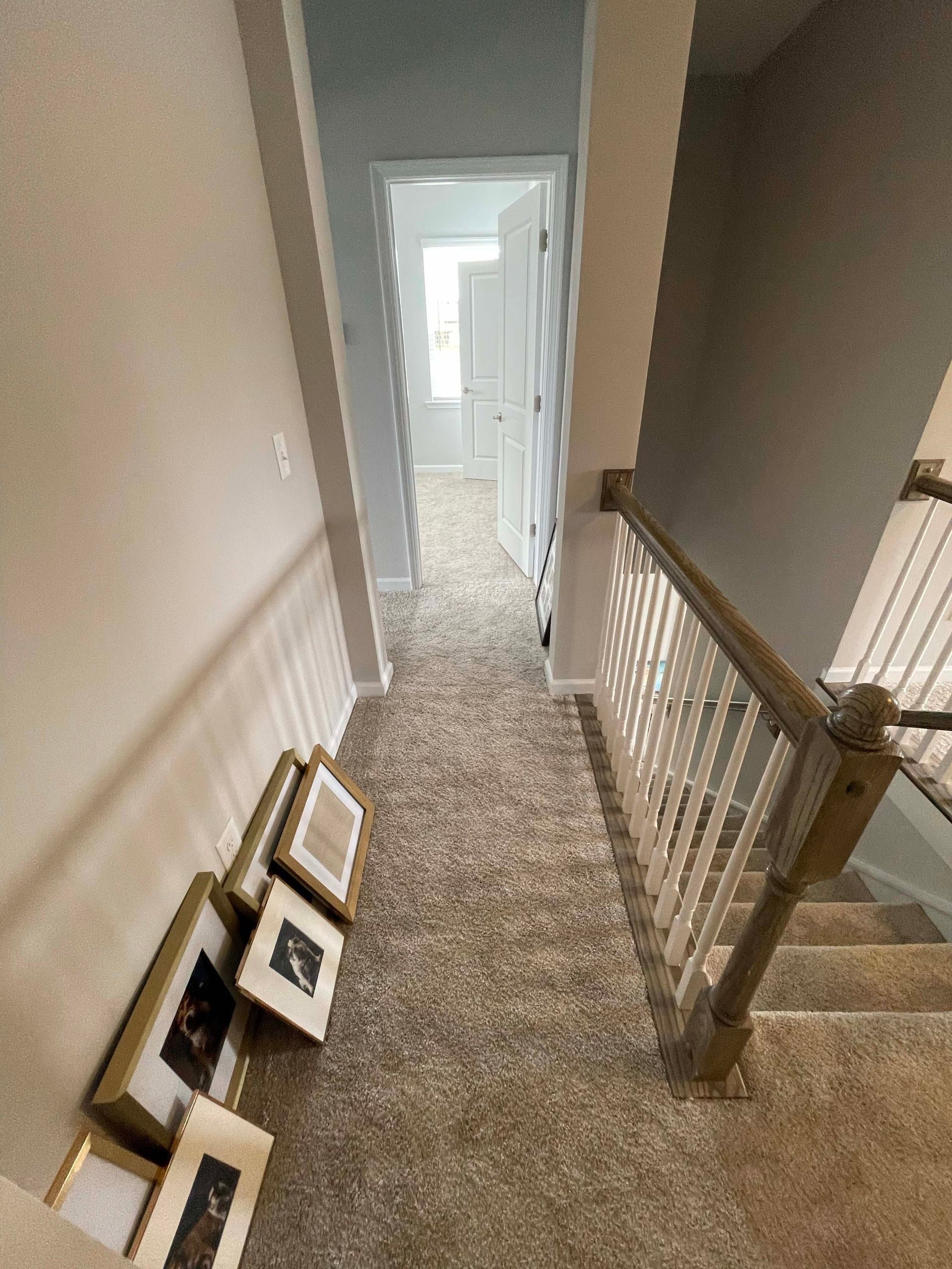Hallway with beige carpet, stairs on right, framed art on the left, door at the end.