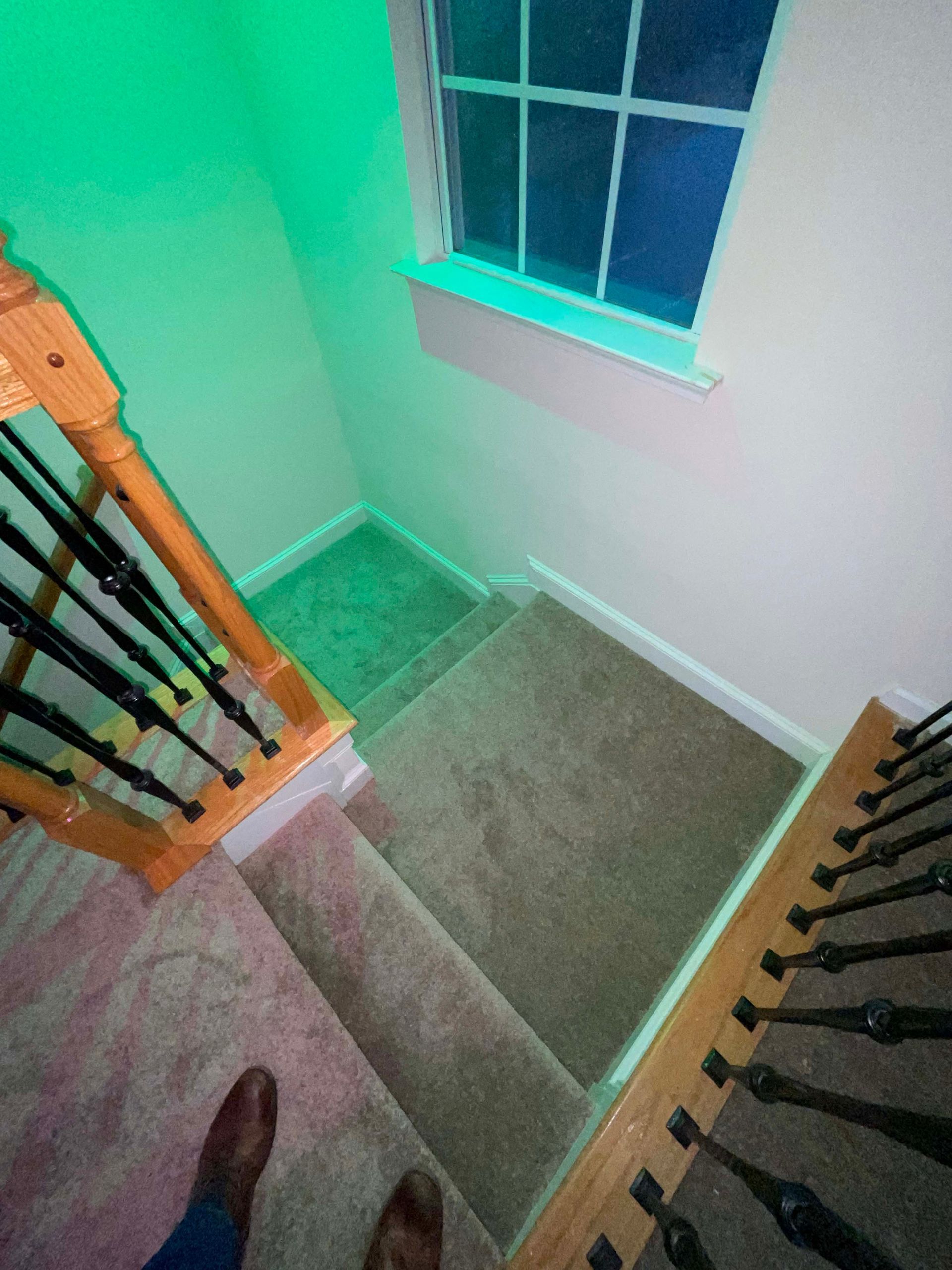 Looking down a carpeted staircase with wood banisters and a window. Green light shines on the wall.