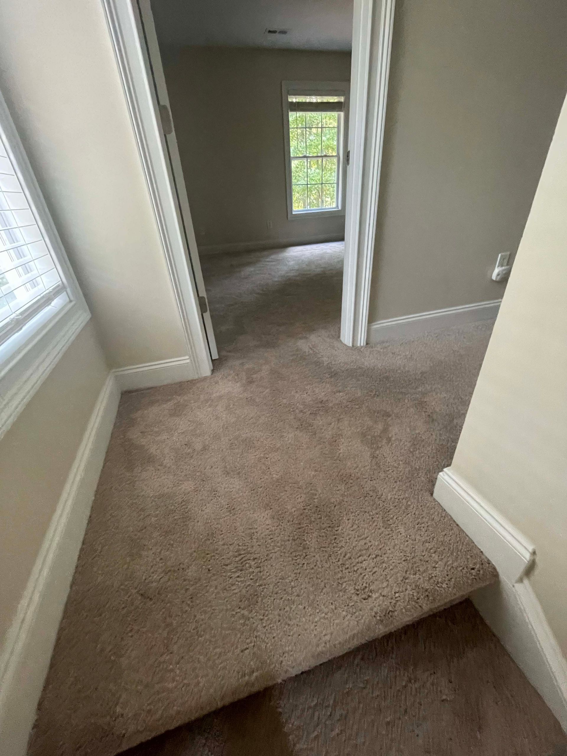 A hallway with carpet, leading to a room with a window. Beige walls and white trim.