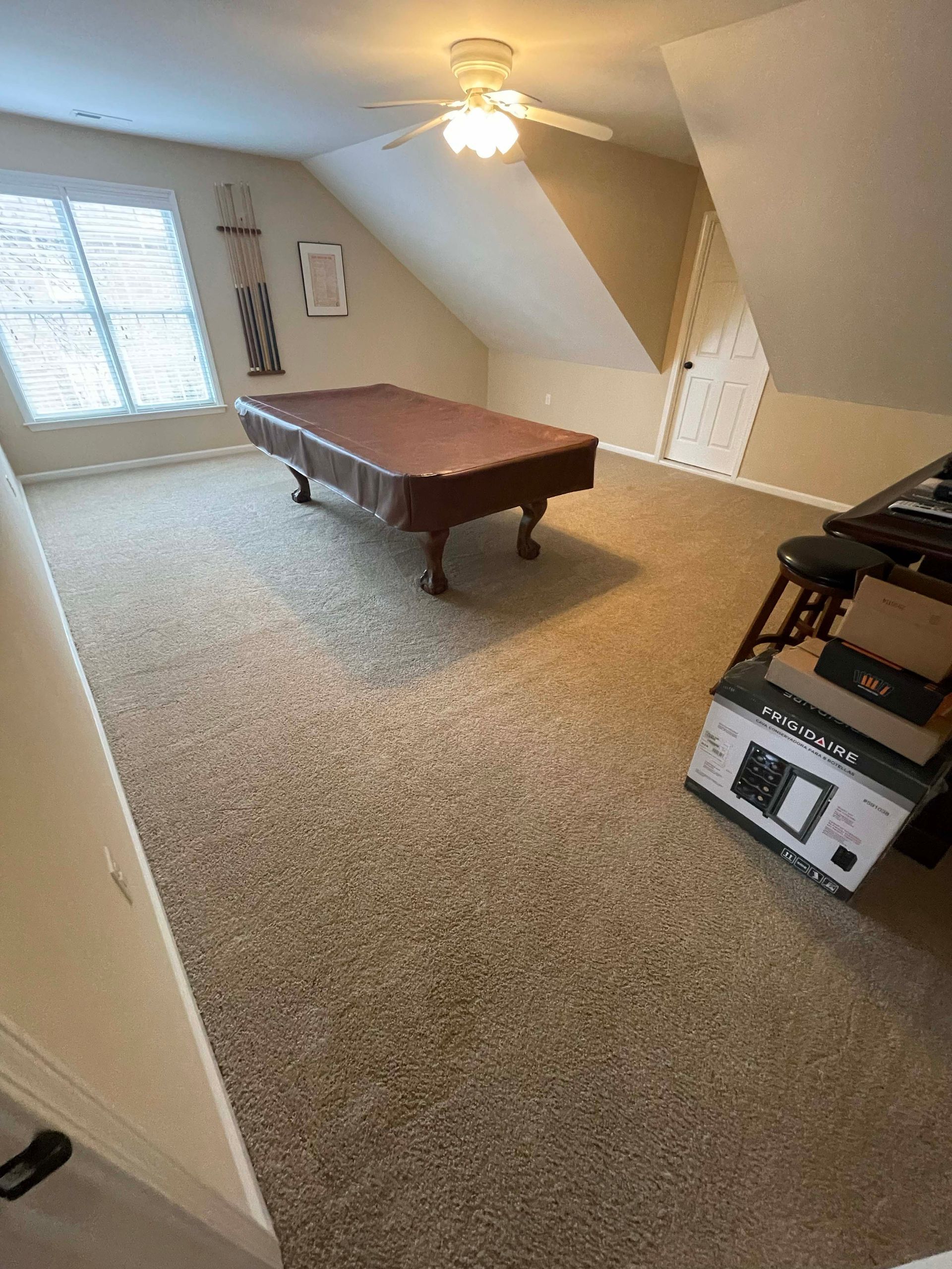 Pool table in a room with angled ceiling and window, brown carpet.