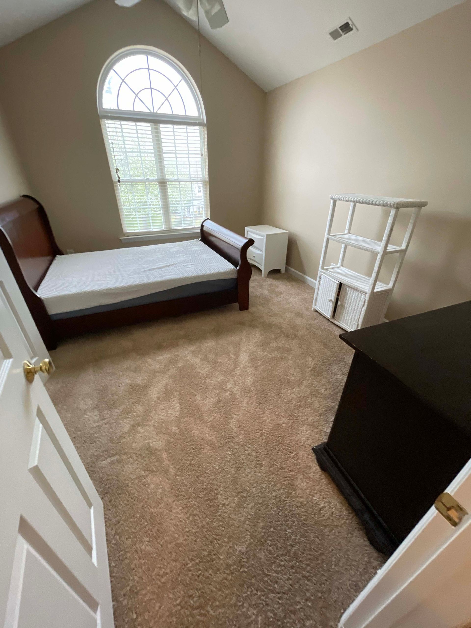 Bedroom with a bed, nightstand, shelf, and dresser; tan carpet and arched window.
