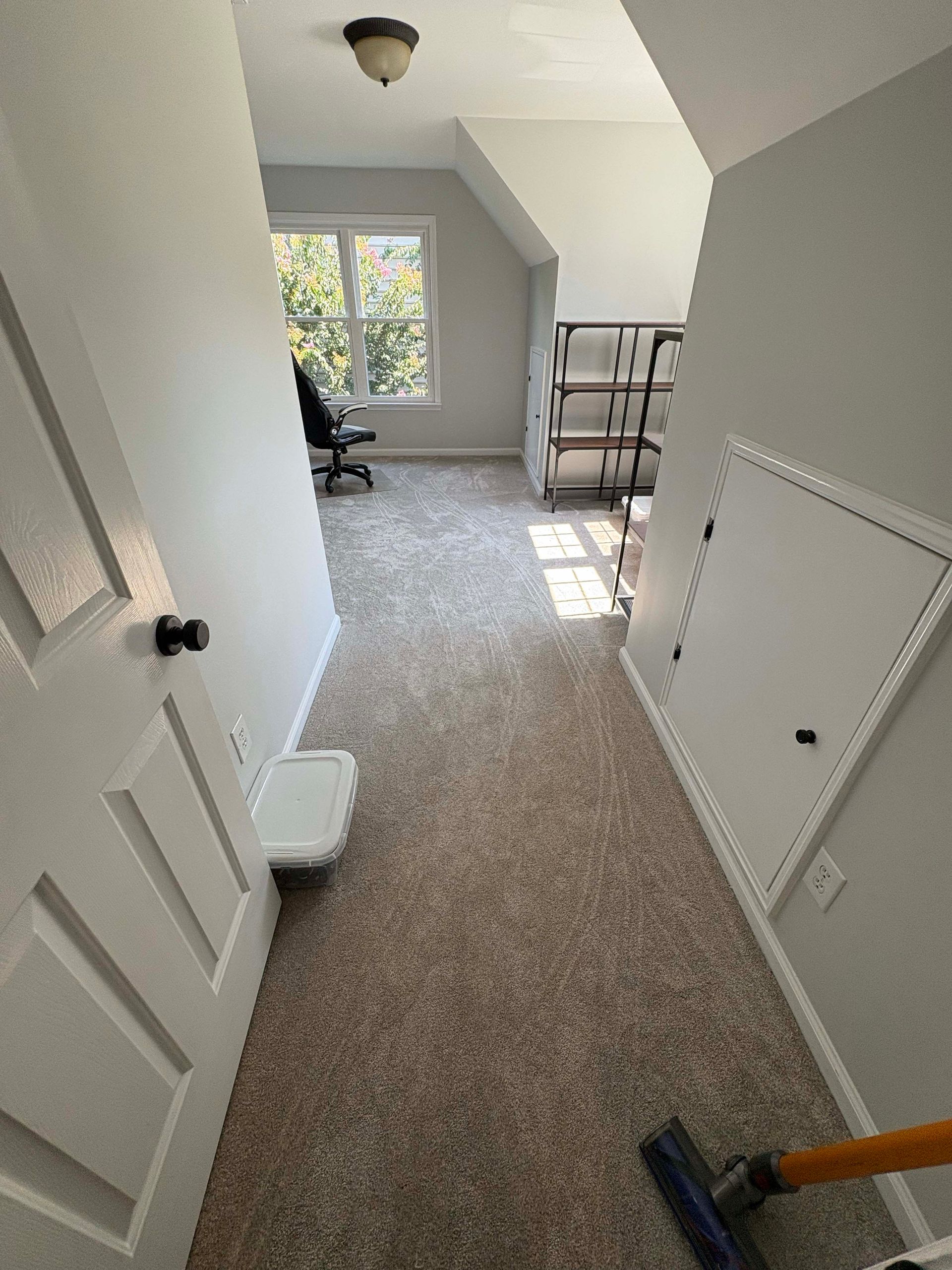 Narrow hallway with beige carpet, white walls, and a door, leading to a room with a window and shelves.