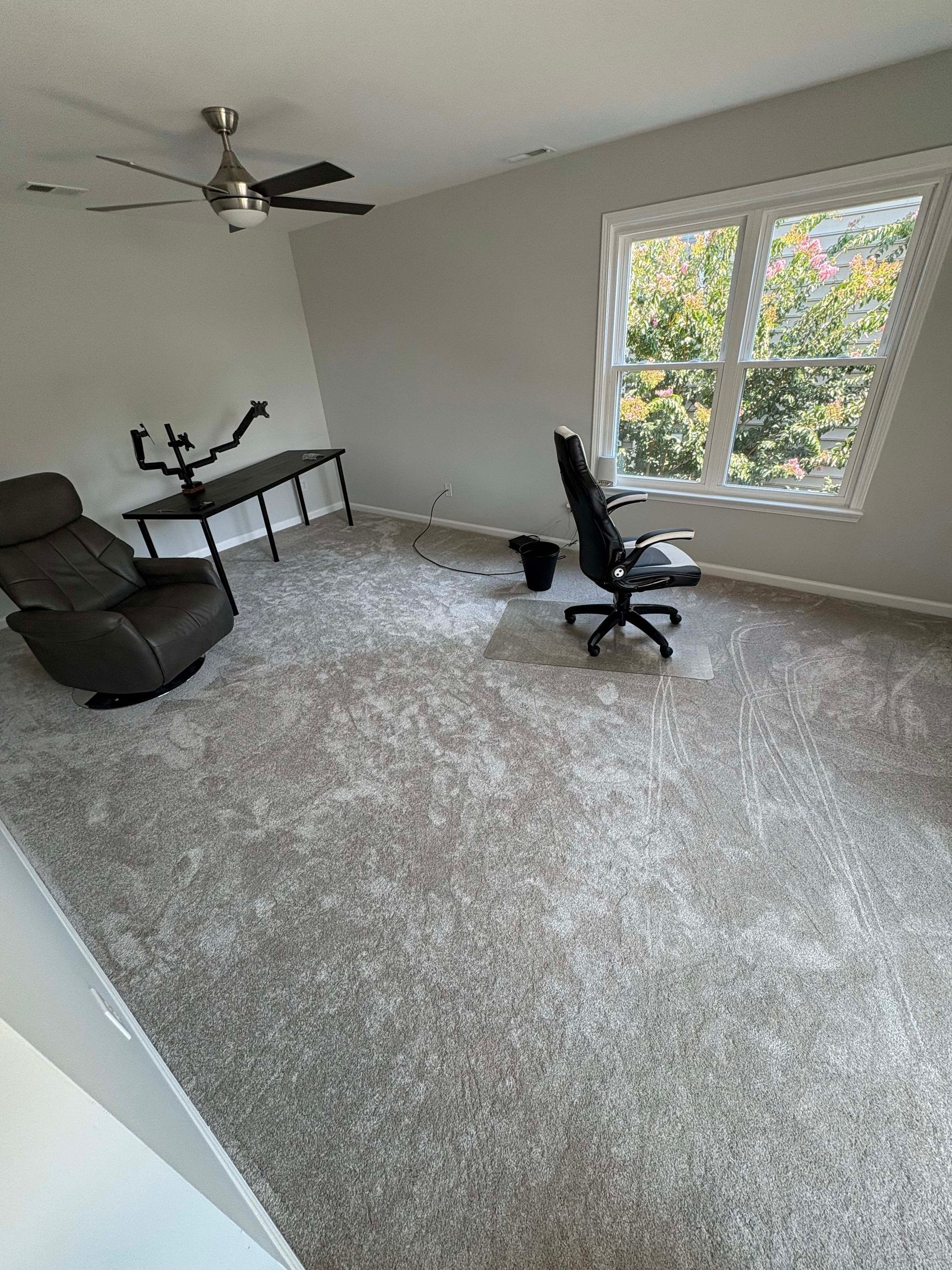 A gray-carpeted room with a reclining chair, desk, and office chair near a window with trees visible.