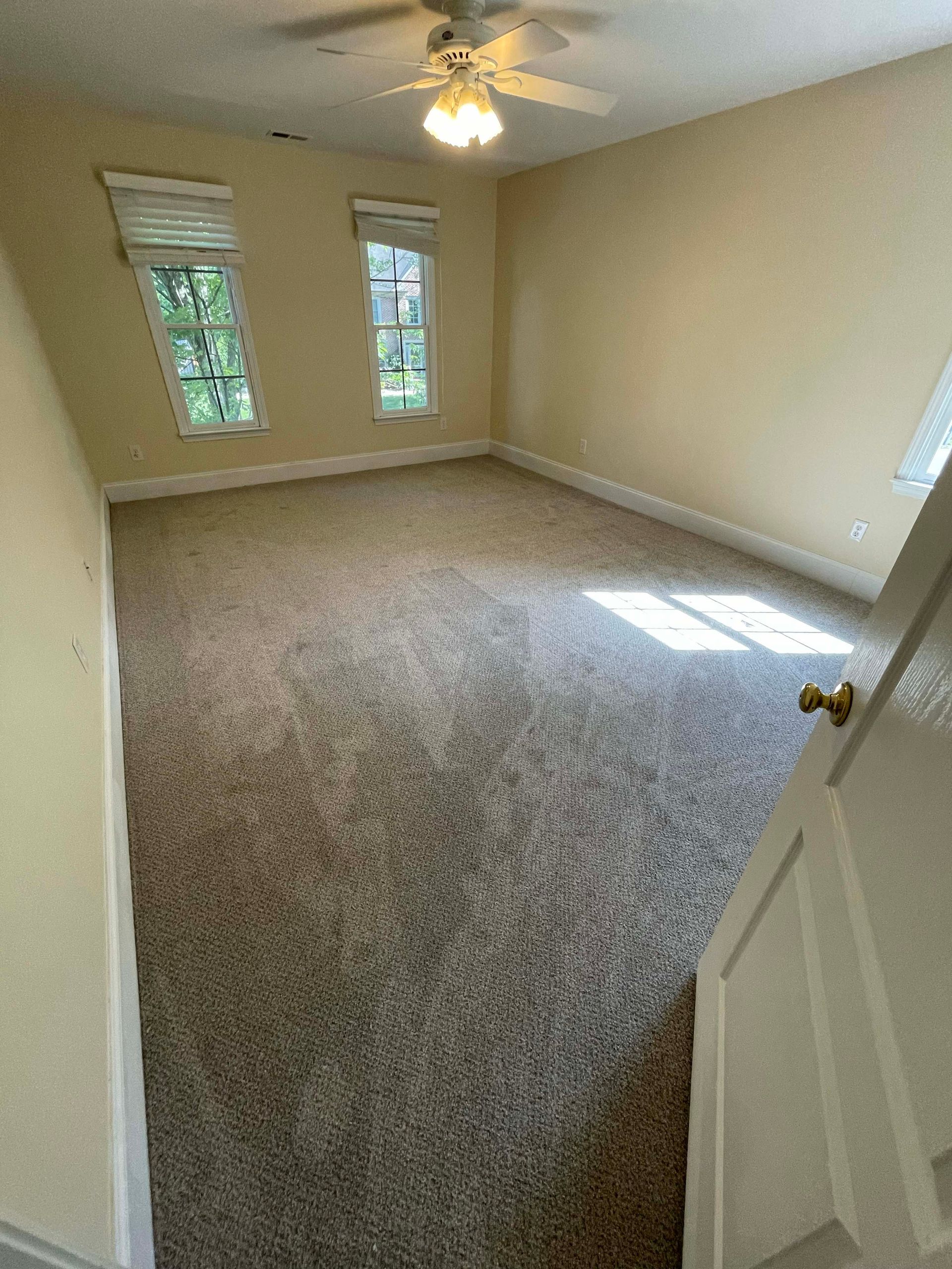 Empty room with beige walls and carpet, two windows, and a ceiling fan.