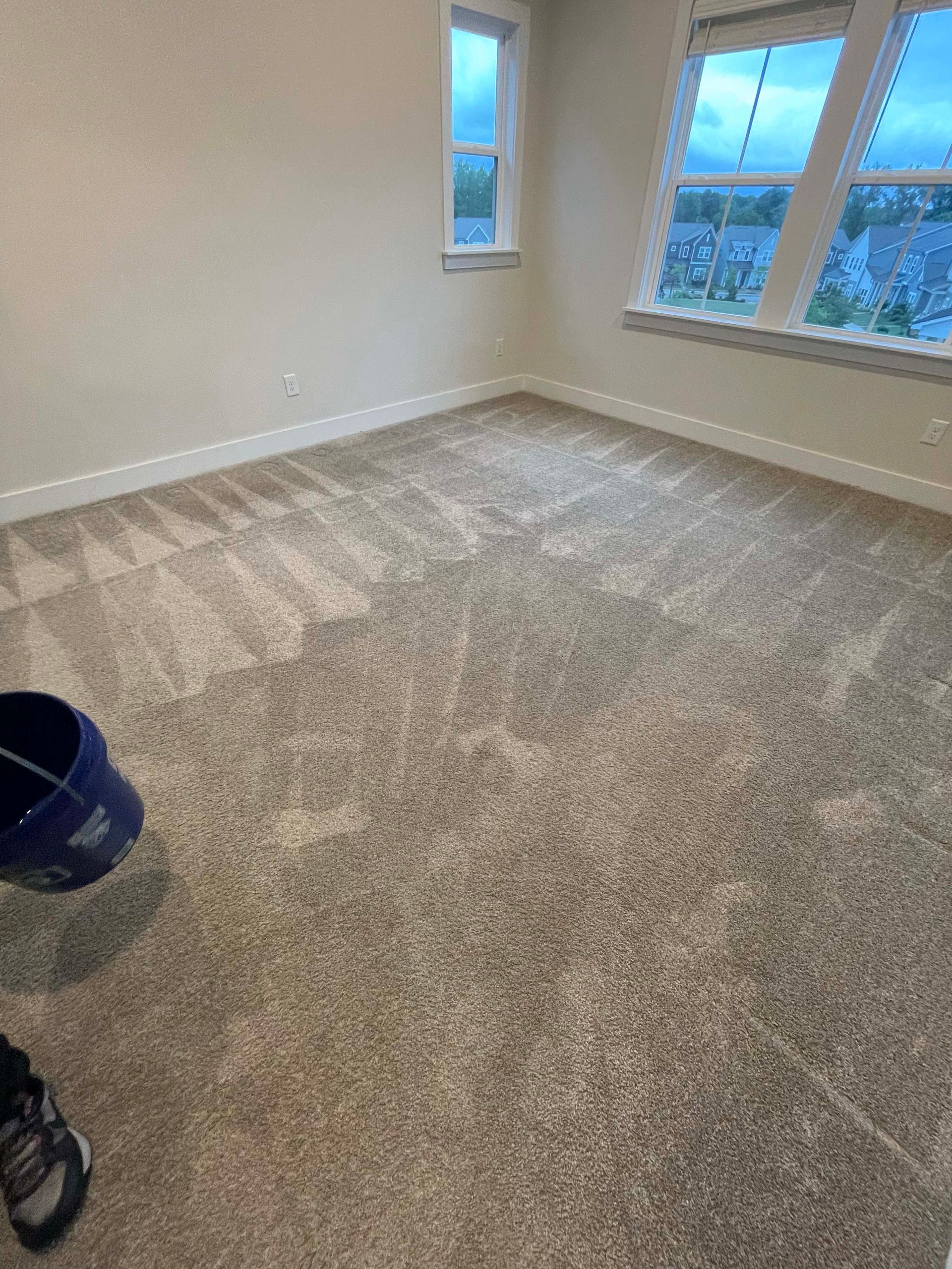 Room with beige carpet, two windows, and a blue bucket.