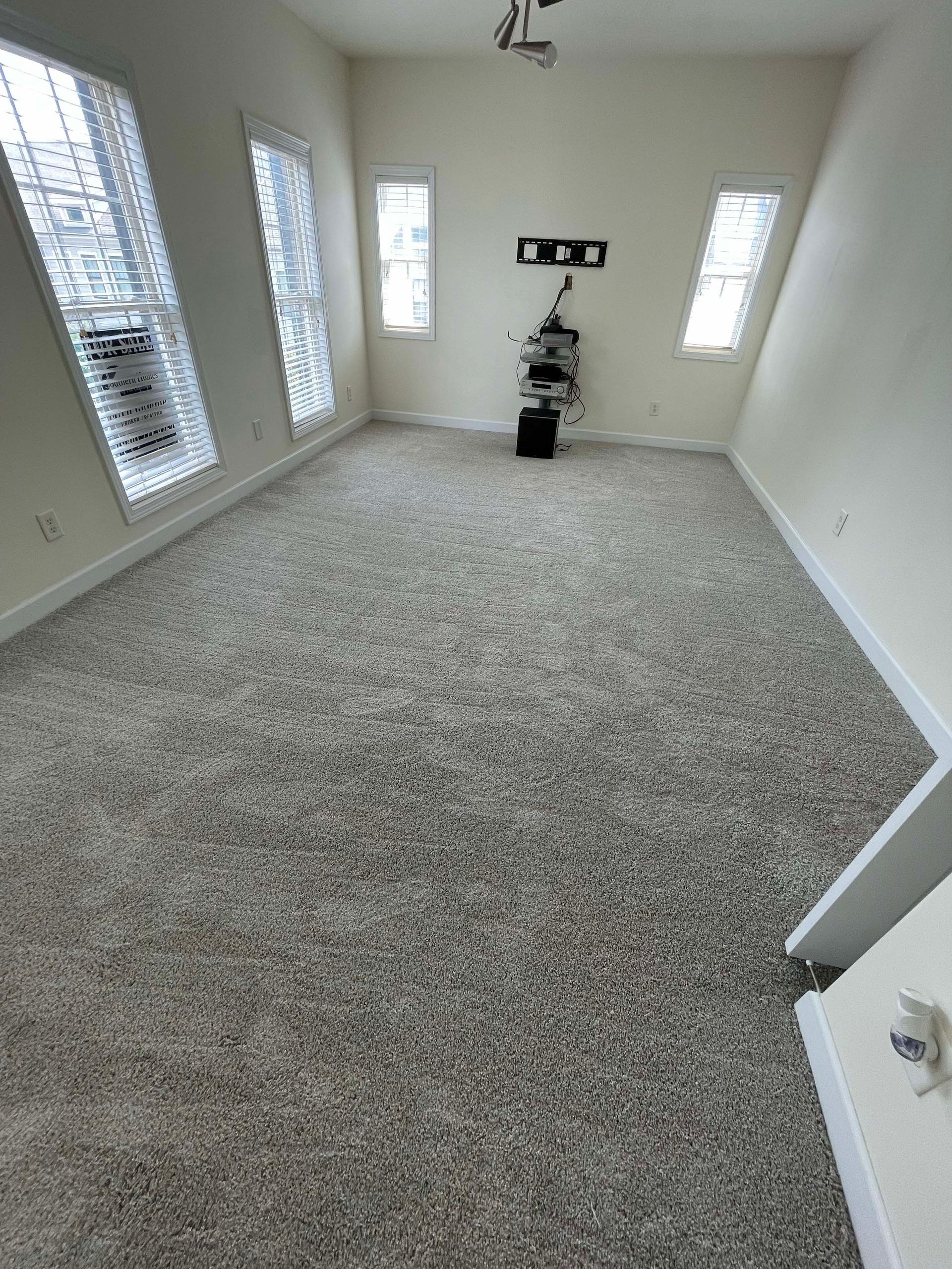 Empty room with light-colored walls, carpet, and windows with blinds. Black television mount and equipment.