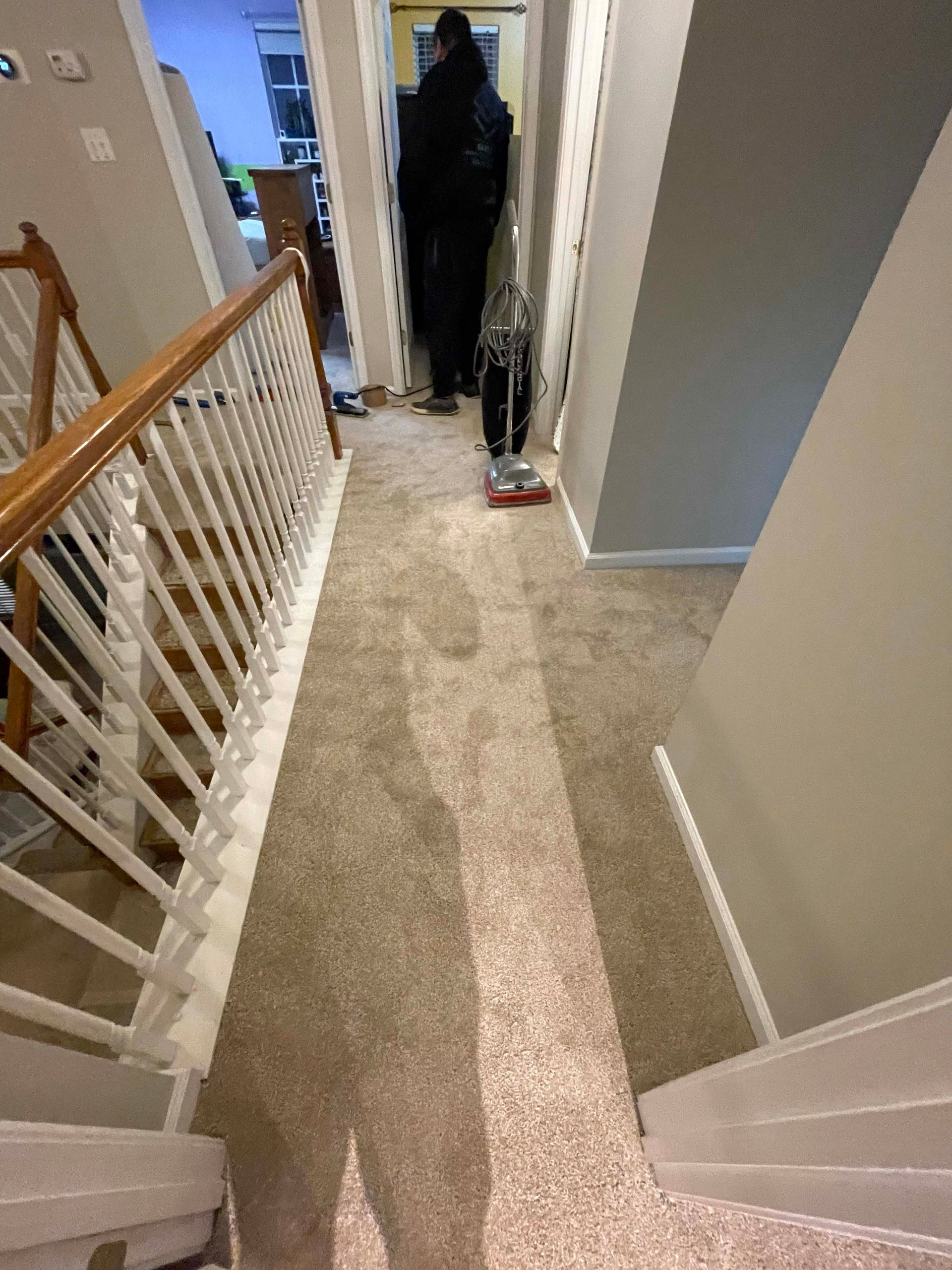 Person vacuums a light-colored carpeted hallway. Stairs are on the left; walls are gray and white.