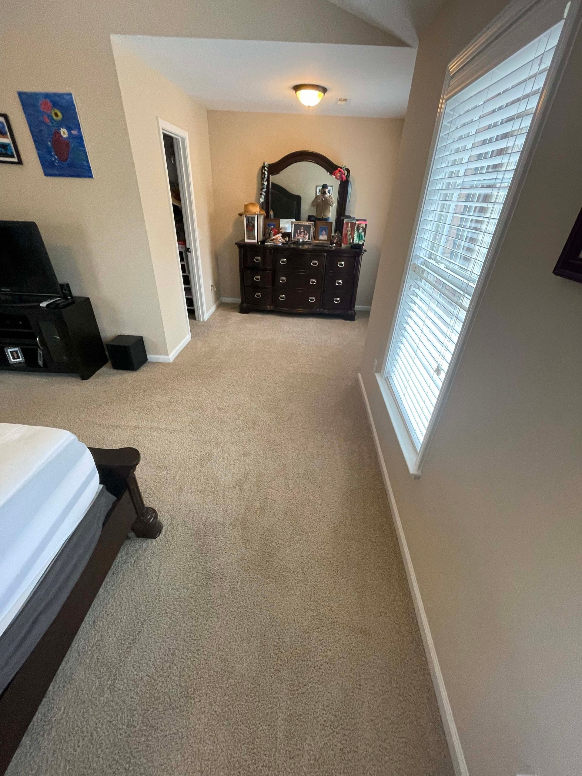Bedroom with bed in foreground, dresser in background, carpeted floor, window with blinds.