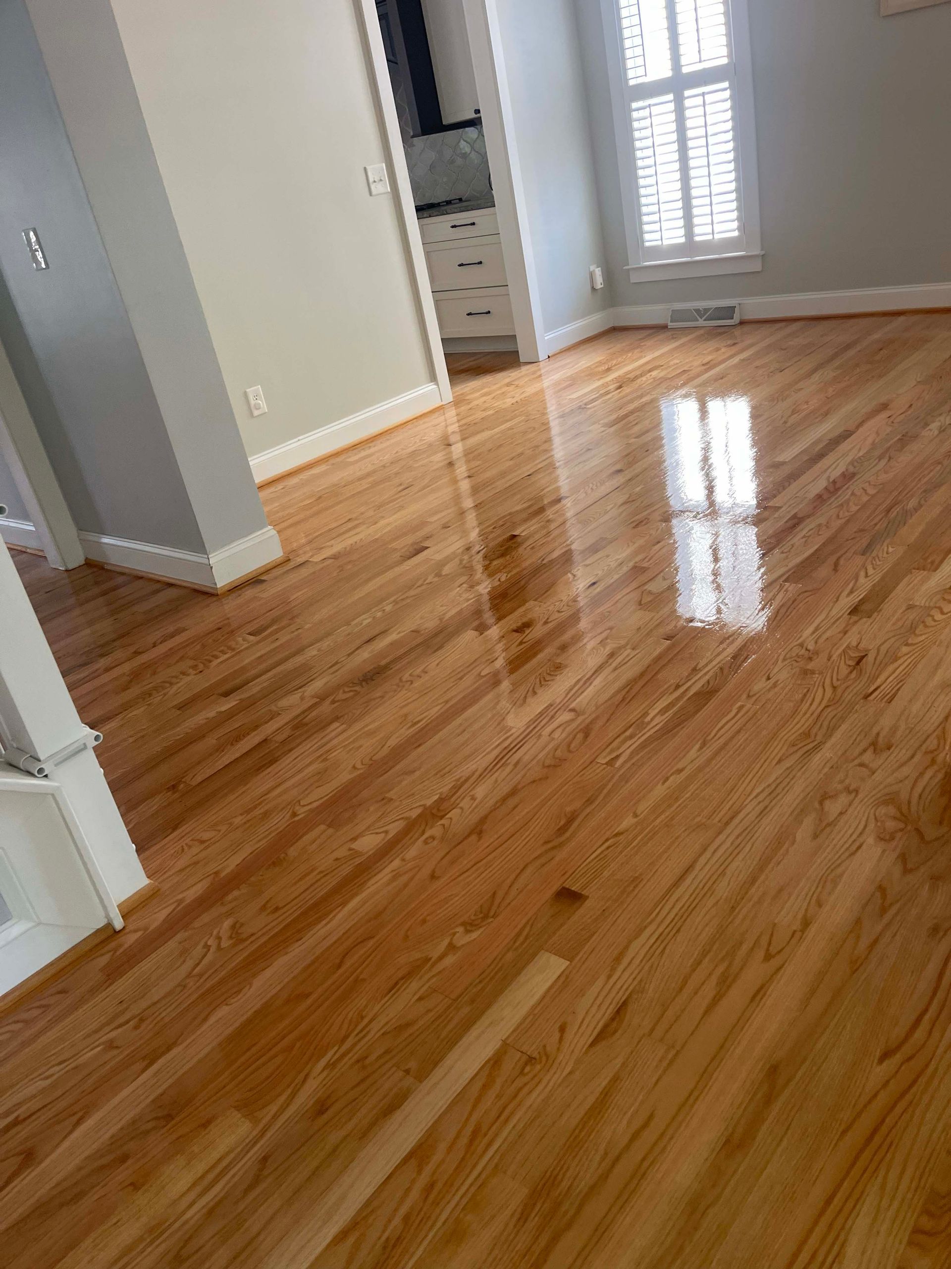 Shiny hardwood floor reflects light from a window in a room with light gray walls.