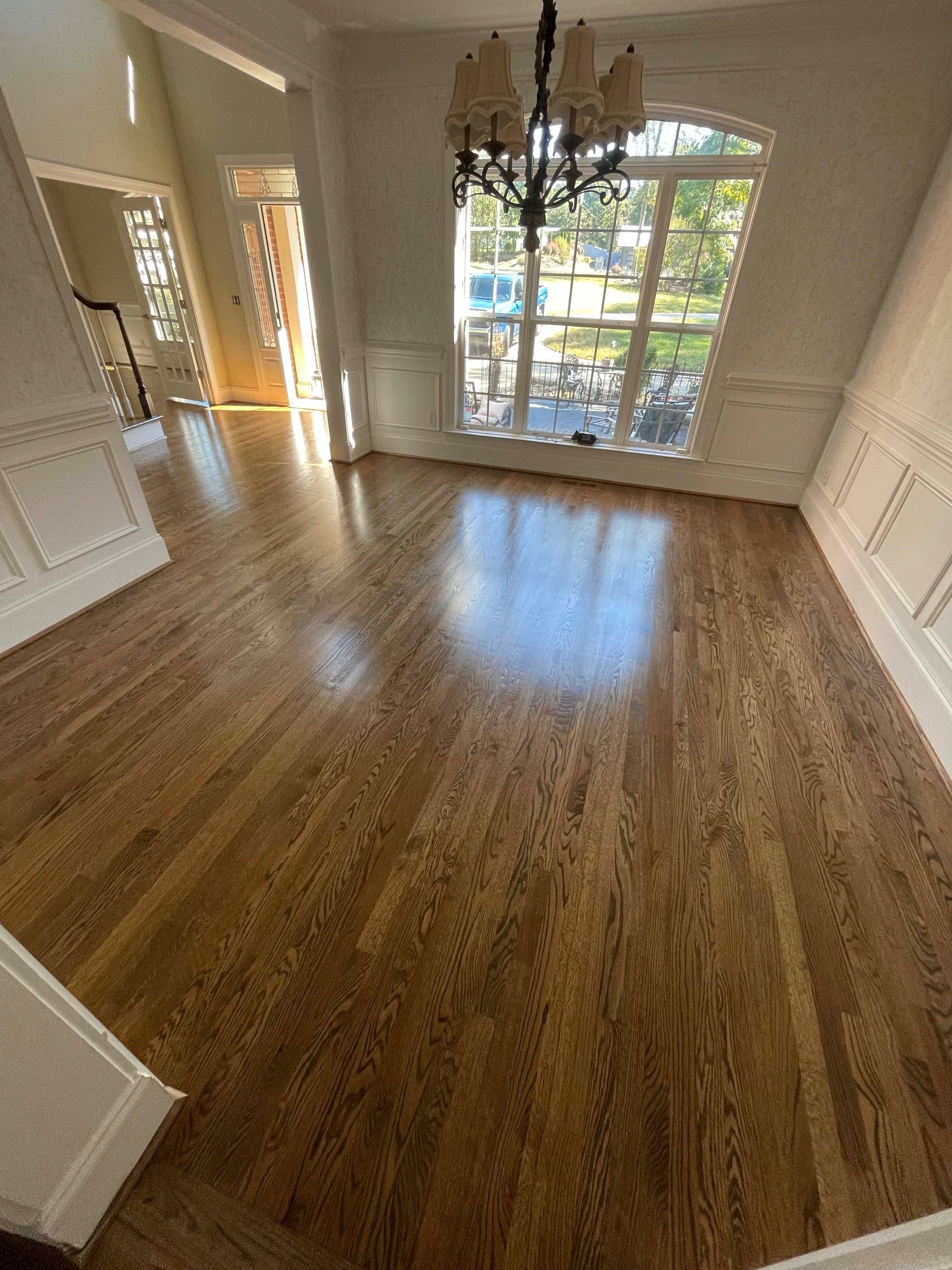 Hardwood floor in a room with white paneling and a large window, chandelier overhead.